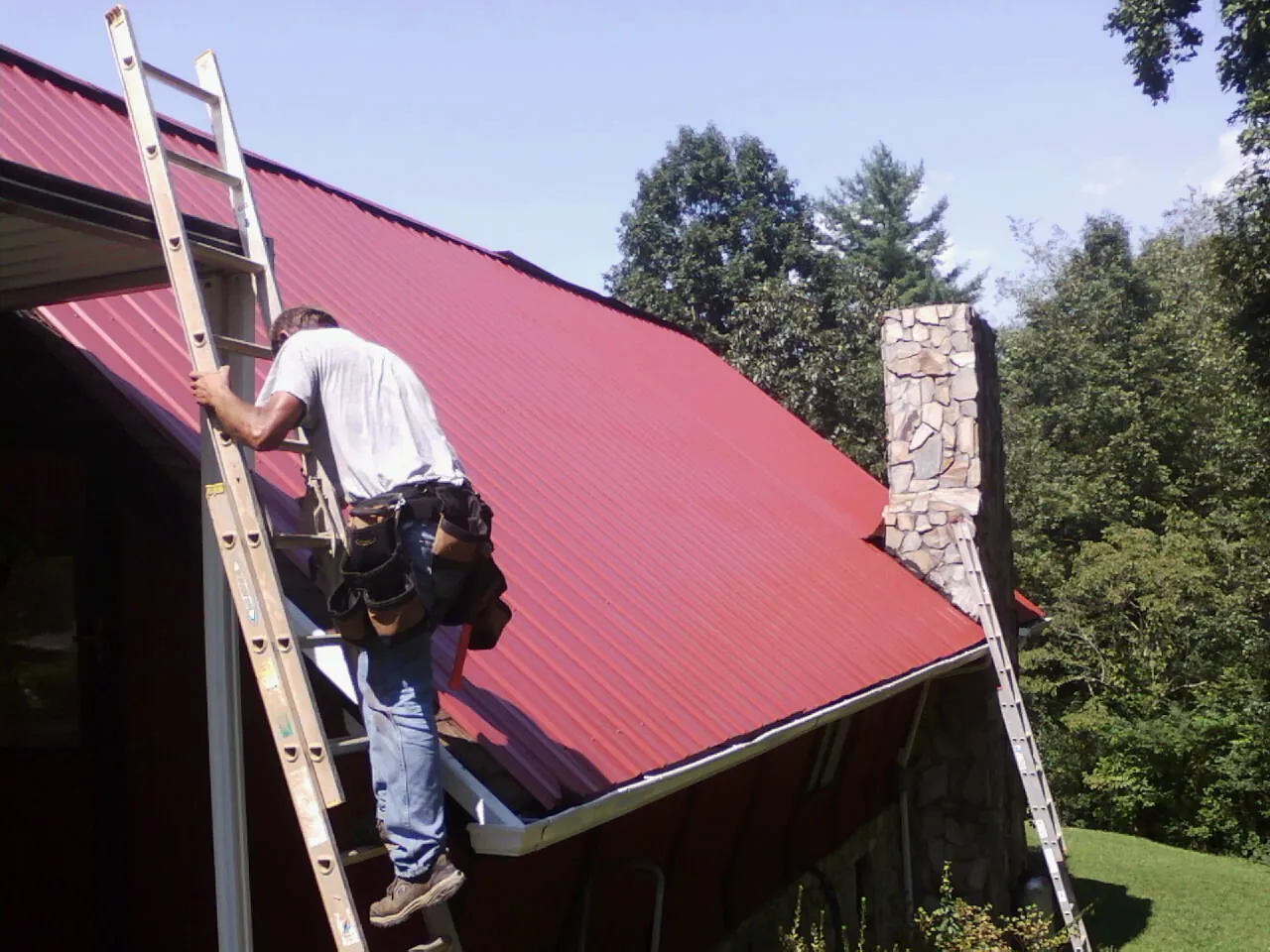 Roofer on a ladder beside a red metal roof and stone chimney — Soffit & Fascia Repair by Roofing Brothers in Cody